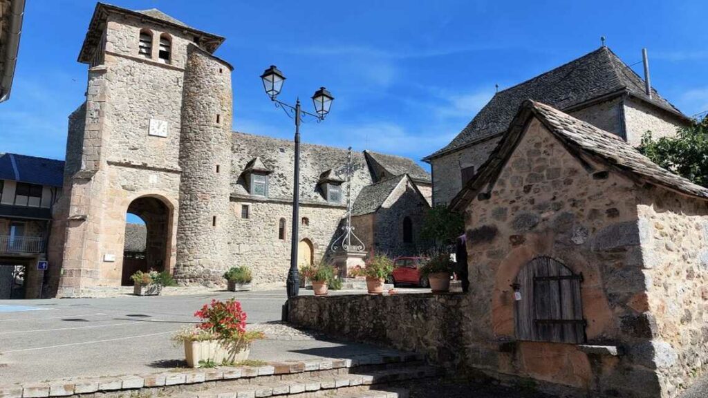 Villeneuve / La Bastide (Aveyron) : porte fortifiée en pierre, maisons aux toits de lauze, lampadaire et ciel bleu.